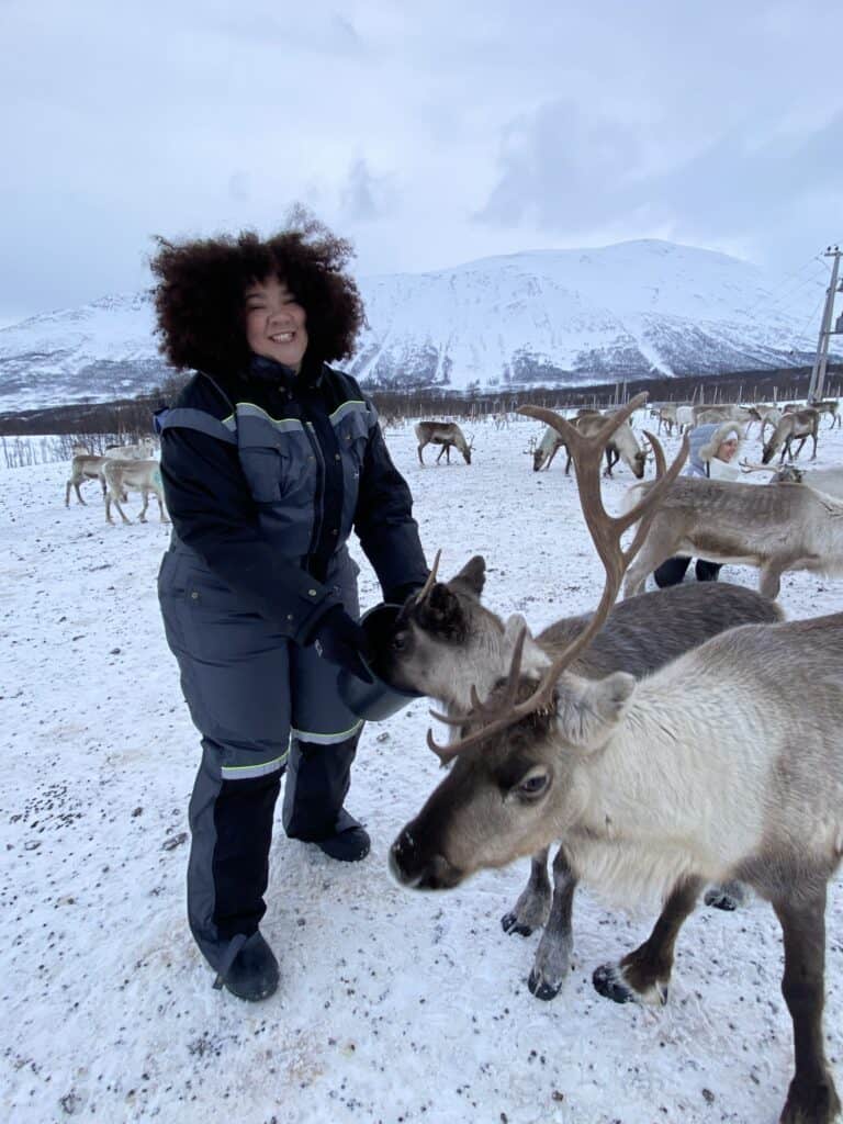 A tall biracial woman is feeding a reindeer from a bucket at a Reindeer farm in Tromso. 