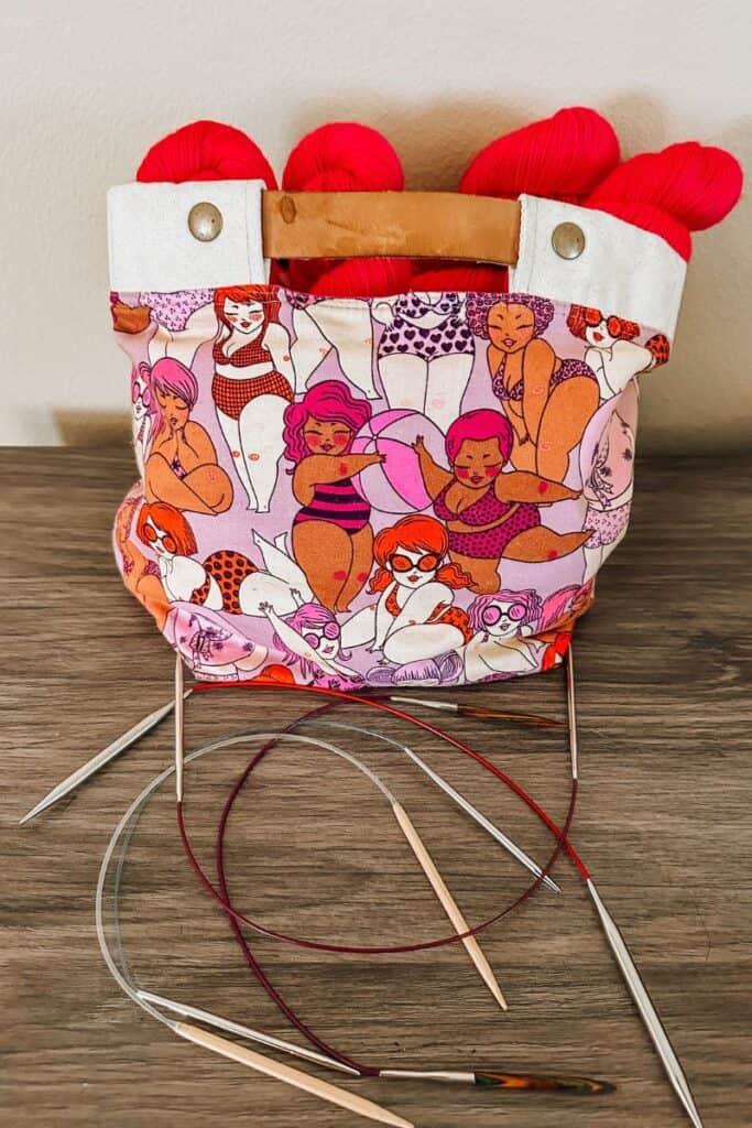 A pink printed knitting bag sitting on a countertop with hot pink yarn sticking it and plastic, metal, and wooden circular needles in front.